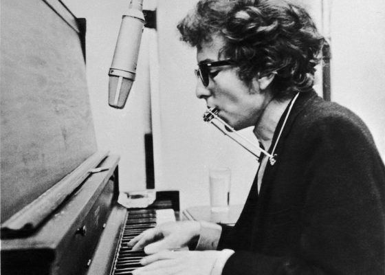 circa 1965: American rock singer and songwriter Bob Dylan playing the piano and the harmonica simultaneously with the use of a harmonica holder. (Photo by Hulton Archive/Getty Images)